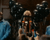 a woman wearing sunglasses is speaking into a microphone in front of balloons