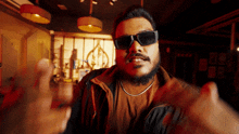 a man wearing sunglasses and a brown jacket is making a gesture