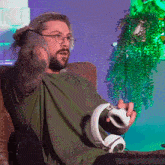 a man with a beard and glasses is sitting on a couch holding a controller and headphones .