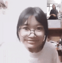 a girl wearing glasses is smiling and looking at the camera while another girl is standing in the background .