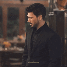 a man in a black suit and black shirt is standing in a room with minimalmoons written below him