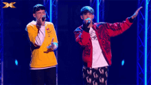 two young men singing into microphones on a stage with an x on the bottom