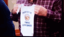 a man in a plaid shirt is holding a white baby onesie that says " property of pawnee government "