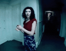 a woman in a red top and blue floral skirt stands in a hallway