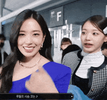 a woman in a blue one shoulder top stands next to another woman in a plaid jacket