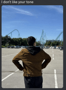 a man in a brown jacket stands in a parking lot looking at a roller coaster in the background