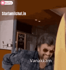 a man wearing sunglasses and a plaid shirt is standing in front of a wall with the words vanakkam written on it .