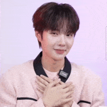 a young man in a pink sweater is smiling with his hands on his chest