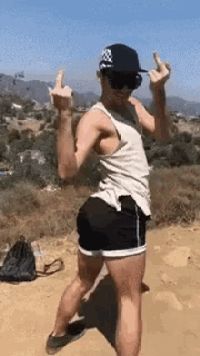 a man in a tank top and shorts is giving the middle finger while standing on a dirt road .