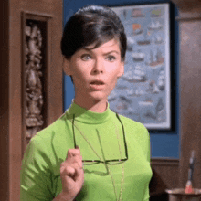 a woman in a green turtleneck is holding a pair of glasses in her hand