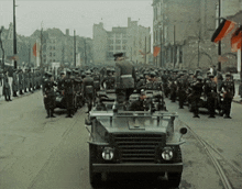a group of soldiers are marching down a street with a jeep in the foreground