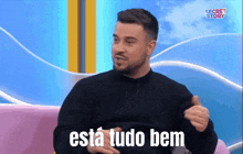 a man with a beard is sitting on a couch with the words " esta tudo bem " above him