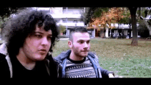 a man with curly hair and a man with a beard are standing next to each other in a park