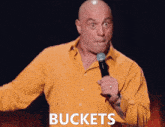a bald man in a yellow shirt is holding a microphone with the word buckets on the bottom right