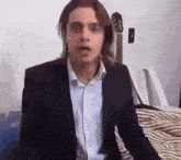 a man in a suit and blue shirt is sitting on a bed and making a funny face .