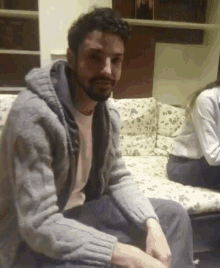 a man with a beard wearing a grey sweater sits on a couch