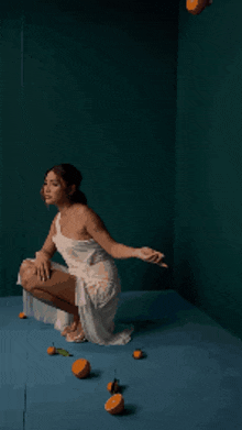 a woman in a white dress is squatting on a blue floor with oranges around her