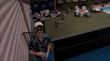 a man wearing sunglasses and a hat stands on a balcony overlooking a lawn