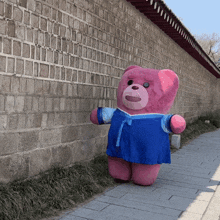 a pink teddy bear wearing a blue dress is standing on a sidewalk in front of a brick wall