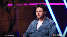 a man wearing a denim shirt is sitting in front of a nbc sign