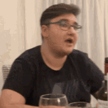a man wearing glasses and a black shirt is sitting at a table with a glass of wine .