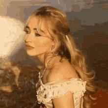 a woman with long blonde hair and a necklace is wearing a white off the shoulder dress .