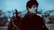 a man in a black coat stands in a field with a man in a helmet in the background