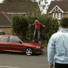 a man in a blue jacket stands in front of a red car while a boy jumps over a fence