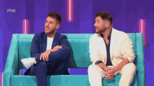 two men sit on a blue couch with rtve written on the bottom right