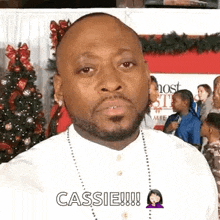 a man with a beard and a necklace is standing in front of a christmas tree and talking to the camera .