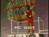 a man in a red jumpsuit is standing on a platform with the words mr. wave behind him