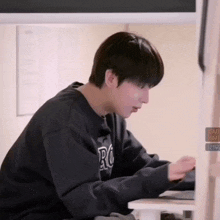 a young man in a black sweatshirt is sitting at a desk with a laptop .
