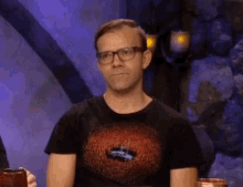 a man wearing glasses and a black shirt is sitting at a table in front of a rock wall .