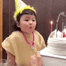 a little girl wearing a party hat is standing in front of a cake with candles .