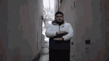 a man in a white jacket is standing in a narrow alleyway with his hands in his pockets .