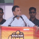 a man is giving a speech in front of microphones and the word khatam is on the bottom