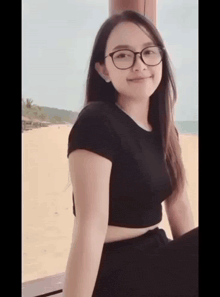 a girl wearing glasses and a black crop top is sitting on a beach .