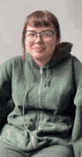 a woman wearing glasses and a green hoodie is sitting in a chair and smiling .
