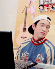 a man wearing a baseball cap is sitting in front of a laptop and a guitar