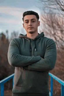 a man in a green hoodie stands with his arms crossed on a bridge