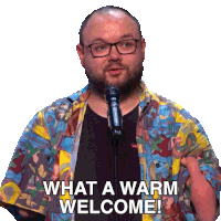 a man wearing glasses and a colorful shirt stands in front of a microphone and says " what a warm welcome "