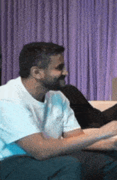 a man with a beard is sitting on a couch in front of purple curtains .
