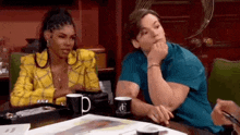 a man and a woman are sitting at a table with cups of coffee . the woman is wearing a yellow jacket .