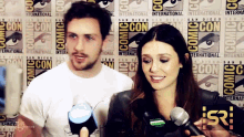 a man and a woman are talking in front of a comic con wall