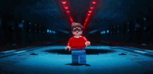 a lego man in a red shirt and blue jeans is standing in a dark room with red lights behind him .