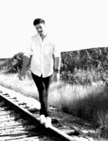 a man in a white shirt and black jeans is walking on train tracks
