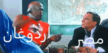 two men are sitting at a table and one is wearing a red shirt and the other is wearing a black suit