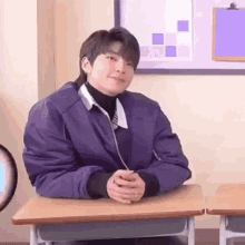 a young man is sitting at a desk in a classroom wearing a purple jacket .