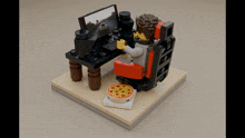 a lego man is sitting at a desk with a computer and a pizza on the floor