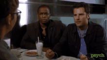 two men are sitting at a table with a drink and a plate of food with the word psych on the bottom right